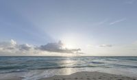 the ocean with a sun behind clouds over the beach and a lone surfboard in the sand