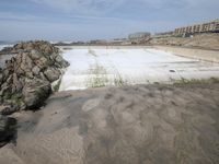 an empty pool of water on the side of a beach near large buildings and a long stretch of sand