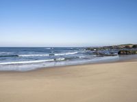 a beach with some waves and rocky shoreline next to it, with a blue sky above