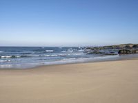 a beach with some waves and rocky shoreline next to it, with a blue sky above