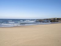 a beach with some waves and rocky shoreline next to it, with a blue sky above