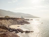 a lighthouse tower is perched at the edge of a mountain and the beach is in the distance