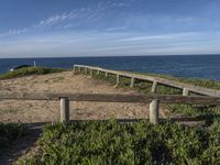 a wooden fence and some plants on a beach with the ocean in the background under a blue sky