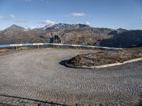 a circular concrete surface in the middle of the road with mountains in the background, on which a metal railing is placed and blue painted