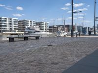 a city street with people walking and sitting by a bench with a boat docked in the background