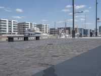 a city street with people walking and sitting by a bench with a boat docked in the background