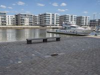 a city street with people walking and sitting by a bench with a boat docked in the background