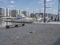 a city street with people walking and sitting by a bench with a boat docked in the background