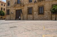 a person walks down a cobbled road past the old building on which a woman is sitting