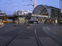 the building that says alexandria plaza in english is next to a train track on a city street