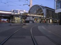 the building that says alexandria plaza in english is next to a train track on a city street