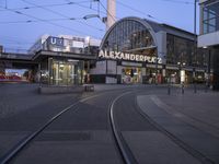 the building that says alexandria plaza in english is next to a train track on a city street