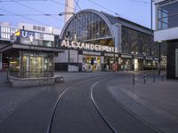 the building that says alexandria plaza in english is next to a train track on a city street