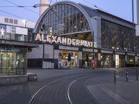 the building that says alexandria plaza in english is next to a train track on a city street