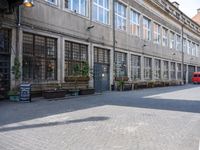 a narrow street lined with tall windows and potted plants next to tables on the street