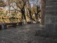 an empty bench sits on a cobblestone street next to some trees and a hill