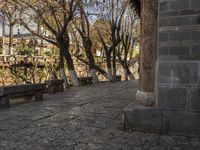 an empty bench sits on a cobblestone street next to some trees and a hill