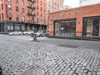 a cobble stone sidewalk with some parked cars and buildings in the background by a street hydrant