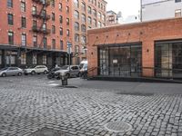 a cobble stone sidewalk with some parked cars and buildings in the background by a street hydrant