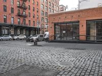 a cobble stone sidewalk with some parked cars and buildings in the background by a street hydrant