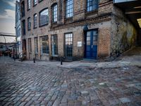 two tall buildings in a narrow alley with cobblestone street below it and a tree in front