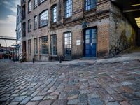 two tall buildings in a narrow alley with cobblestone street below it and a tree in front