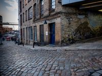 two tall buildings in a narrow alley with cobblestone street below it and a tree in front