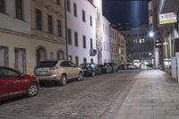 a cobblestone street lined with parked cars at night time, at an intersection in a small european city