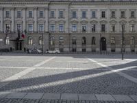 a large building with two people walking in the middle and one car driving on a cobblestone street