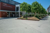 an empty parking lot with people coming out of it and some bushes next to the building