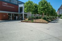 an empty parking lot with people coming out of it and some bushes next to the building