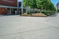 an empty parking lot with people coming out of it and some bushes next to the building