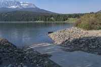 the mountains are covered with snow and water behind a concrete bench near a lake with rocks on a cement shore