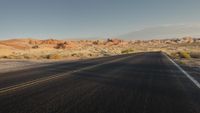 the empty road is lined with desert bushes and sand dunes as the sun sets on the horizon