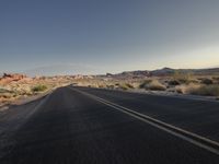 the empty road is lined with desert bushes and sand dunes as the sun sets on the horizon