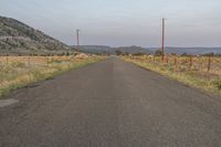 the end of a road that is almost empty on it's side in a wide open field, with telephone poles on the other side and distant hills in the distance