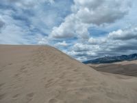 sand dunes and mountains under a cloudy blue sky with clouds in the distance taken from behind it