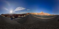 a panorama of a desert road with hills in the distance and a person riding down a curved road