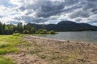 a beach next to mountains in the background with grass and trees lining the shore of the river