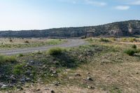 a dirt road surrounded by dry grass and rocks with a dirt field behind it with hills in the background