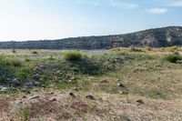 a dirt road surrounded by dry grass and rocks with a dirt field behind it with hills in the background