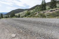 the asphalt in the middle of a gravel road with hills behind it and pine trees along the side