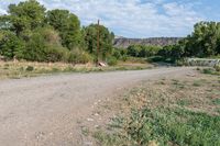 an empty gravel road with dirt and bushes at the side of it and a rusty sign in the background