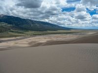 sand dunes with mountains, clouds and a stream in the distance across the landscape with trees