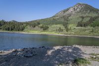 a mountain rises behind a body of water near a dirt shore and some trees and rocks