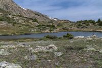 a green meadow near the mountains with a body of water under it and lots of rocks