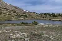 a green meadow near the mountains with a body of water under it and lots of rocks