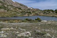 a green meadow near the mountains with a body of water under it and lots of rocks