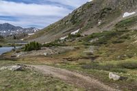 a dog is standing on the side of a dirt trail overlooking mountains with lake and grass