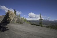 a mountain side road with a large rock cliff in the background as clouds move across the blue sky